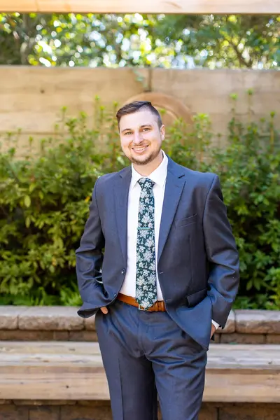 Kyle Ames pharmacist, man in a suit, standing in front of a bench and foliage.